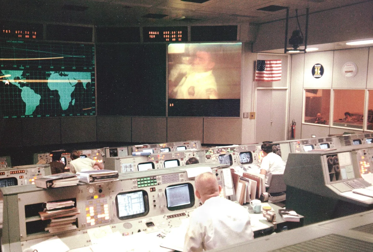 La salle de contrôle de mission de la NASA pendant Apollo 13, en 1970 : des rangées de consoles et d’écrans, des ingénieurs debout.