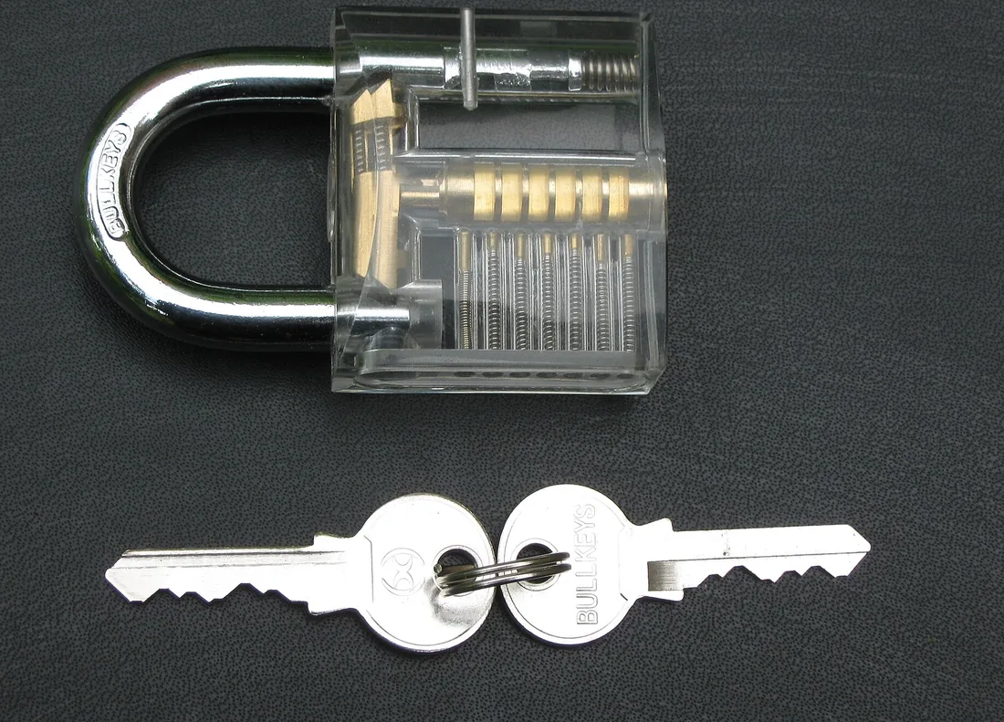 Un cadenas à corps transparent, mécanisme visible, posé à côté de deux clés sur un fond sombre.